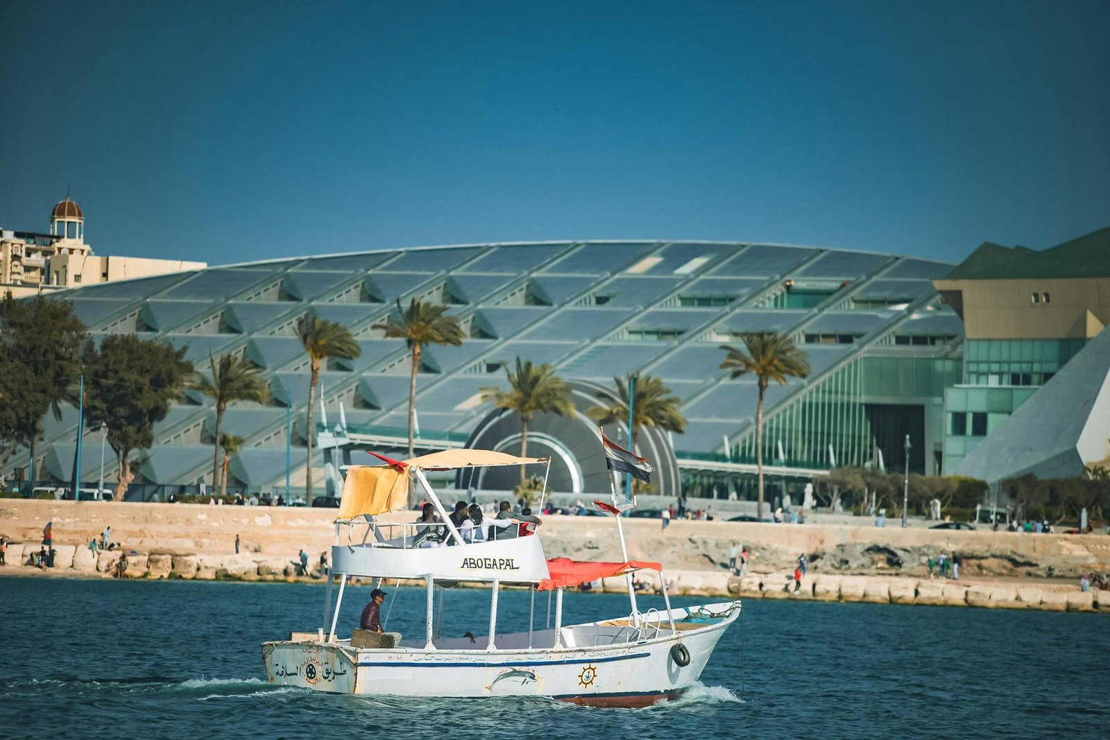 A leisure boat sailing by the Bibliotheca Alexandrina with a backdrop of palm trees, showcasing Alexandria's vibrant seascape.